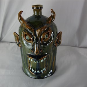May include: A ceramic jug with a devil face design. The jug is primarily green with brown accents on the horns, ears, and facial features. The devil's face has a menacing expression with sharp teeth and piercing blue eyes.