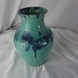 May include: A ceramic vase with a turquoise glaze and dark blue drip pattern. The vase has a rounded body and a flared rim. The handmade pottery is a decorative item.