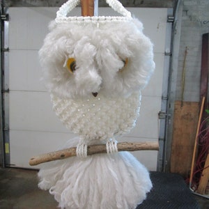 May include: White crocheted owl wall hanging with yellow eyes and a wooden branch. The owl is hanging from a wooden post.