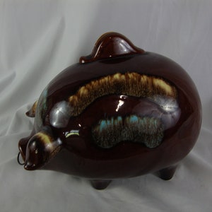 May include: A brown ceramic piggy bank with a glazed finish. The glaze has brown, yellow, and blue swirls. The piggy bank has a metal coin slot on the side.
