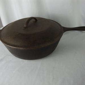 May include: A dark brown cast iron skillet with a lid. The skillet has a long handle and a loop handle on the lid. The cookware is made of cast iron and is designed for cooking. The skillet is sitting on a white surface.