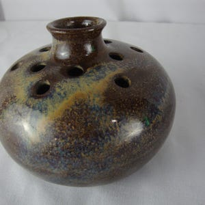 May include: A round, brown ceramic flower frog vase with multiple holes on the top and sides. The vase has a speckled glaze with blue and yellow accents. The vase has a small, cylindrical neck.