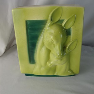 May include: A rectangular ceramic vase in a light lime green glaze featuring a raised relief design of two deer. The relief shows one larger deer standing behind a smaller deer, with a dark green rectangular accent panel behind the figures.