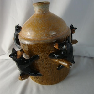 May include: A ceramic jug with a textured brown finish resembling tree bark. The vessel features three small, glossy black bear figurines sculpted in relief, appearing to climb up the sides of the jug toward the narrow, rounded neck opening.