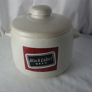 May include: A cream-colored ceramic crock with a matching lid and two side handles. The front features a rectangular red label with a black border containing the white text Black Label BEER. The ceramic has a speckled texture throughout.