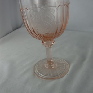 May include: A pink-tinted glass goblet with a detailed floral pattern etched into the bowl. The stem and base are also pink, with a circular, ridged base. The glass has a vintage aesthetic.