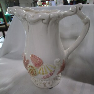 May include: White ceramic pitcher with a seashell design. The pitcher has a wide mouth and a handle that curves up and over the top. The seashells are painted in shades of pink, yellow, and green.