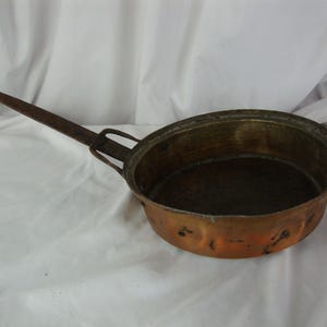 May include: A round copper pan with a dark, weathered patina and a long, slender iron handle. The pan features a deep, circular basin with visible signs of wear and oxidation on the exterior surface, attached to the handle by a metal bracket.
