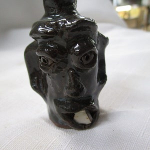 May include: A small, black ceramic figurine of a face with a wide mouth and large eyes. The figurine is glazed and has a shiny finish.