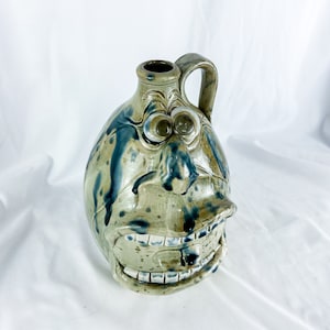 May include: A ceramic pitcher with a whimsical face. The pitcher is glazed in shades of green and blue with brown accents. The face has large eyes, a wide mouth, and a long nose.