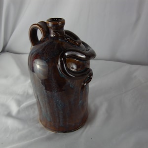 May include: A brown ceramic jug with a glossy finish. The jug has a handle and a decorative, looping design around the neck. The glaze has blue and brown tones.