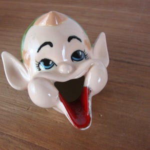 May include: A vintage ceramic ashtray shaped like a smiling elf face. The elf has blue eyes, a red tongue, and a peach-colored face with green and tan hair. The ashtray is on a wooden surface.