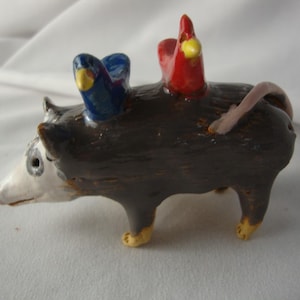 May include: A ceramic figurine of a dark brown opossum with a white face and yellow-tipped feet. Perched on the back of the opossum are two small birds, one glossy blue and one glossy red, both featuring yellow beaks and black eyes.