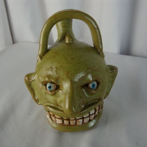 May include: A green ceramic pitcher shaped like a goblin head with a handle on top. The vessel features a wide, toothy grin, prominent ears, and two bright blue eyes set into the face. The glaze has a mottled, earthy texture throughout.