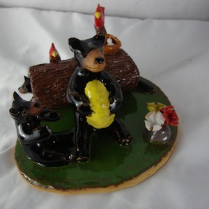 May include: A ceramic figurine featuring black bears on a green base. One bear stands holding a yellow object, while another reclines. A brown log and small flowers are also present. The figurine has a glossy finish.