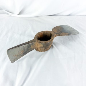 May include: A vintage pickaxe with a wooden handle and two metal blades. The pickaxe has a weathered appearance, with signs of rust and wear on the metal. The blades are a dull silver color, and the handle is a dark brown.