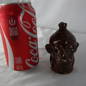 May include: A brown ceramic jug in the shape of a face with a smiling expression. Next to it is a red Coca-Cola can with white lettering and a QR code. The can is 12 fl oz (355 mL).
