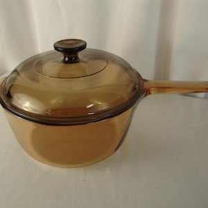 May include: A transparent amber-colored glass saucepan with a matching lid. The pot features a long, integrated glass handle on one side and a small tab handle on the opposite side. The circular lid includes a central knob for easy removal.