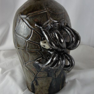 May include: A ceramic vase with a spider design. The vase is brown and black with a spider web pattern. The spider has eight legs and is made of black clay.