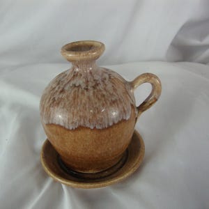 May include: A small, round ceramic jug with a handle and a matching saucer. The jug is brown with a lighter, textured top. The saucer is also brown. The jug and saucer are likely for decorative purposes.
