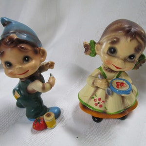 May include: Two ceramic figurines of children. The boy is wearing a blue hat and green overalls and holding a needle and thread. The girl is wearing a yellow dress with red flowers and holding a blue bowl with red strawberries.