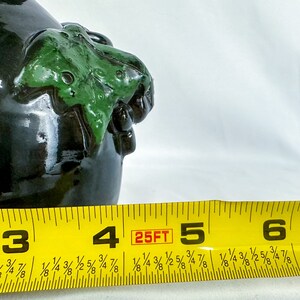 Puede incluir: Primer plano de una botella de cer&aacute;mica verde oscuro con un dise&ntilde;o esculpido verde y marr&oacute;n. Una cinta m&eacute;trica amarilla con marcas negras indica "25FT" y muestra los n&uacute;meros 3, 4, 5 y 6.