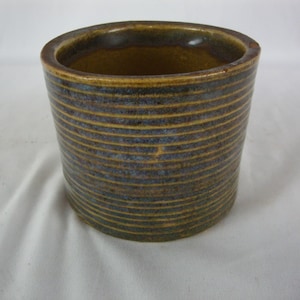 May include: A cylindrical ceramic container with horizontal bands of yellow and blue-grey. The interior is a darker brown. The rim is rounded and the exterior has a textured appearance. The container is likely a small planter or a decorative item.