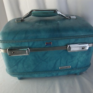 May include: A teal hardshell train case with a textured finish, featuring a chrome-colored metal handle and latch hardware. The front displays a small rectangular logo plate with the text Tourister in white lettering on a dark background.