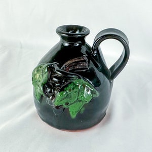 Puede incluir: Una peque&ntilde;a jarra de cer&aacute;mica negra con un dise&ntilde;o de hojas verdes y marrones. La jarra tiene un asa y un cuerpo redondeado.