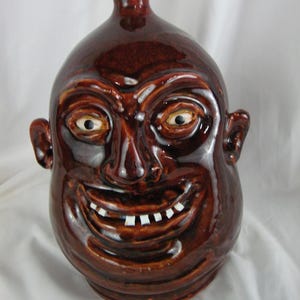 May include: A brown ceramic jug shaped like a face with a wide smile and white teeth. The jug has a brown glaze and a small opening at the top.