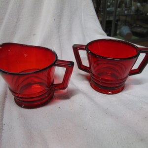 May include: A set of two red glass creamer and sugar bowls with a ribbed design.