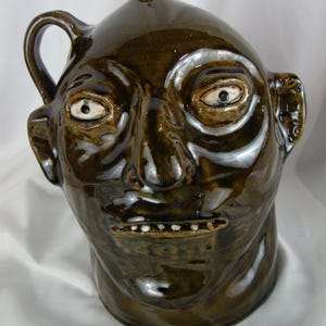 May include: A brown ceramic jug shaped like a human face. The jug has a handle on one side and a small spout on top. The face has detailed features, including eyes, a nose, and a mouth with small teeth. The jug is glazed with a glossy finish.