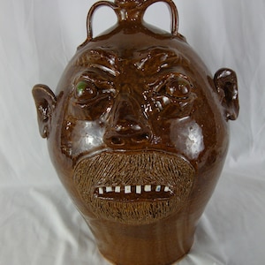 May include: A brown ceramic jug with a face sculpted on the front. The face has a wide mouth with white teeth, a bushy beard, and large, green eyes.