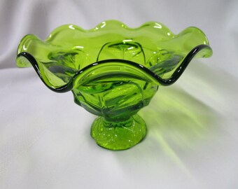 May include: A green glass bowl with a ruffled rim and a pedestal base. The bowl has a textured surface and is perfect for serving fruit or snacks.