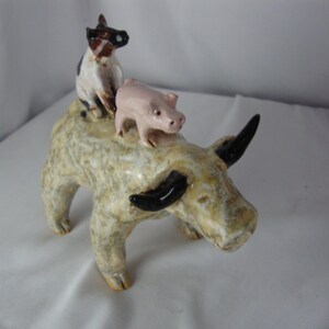May include: A ceramic sculpture of a bull with a dog and a pig riding on its back. The bull is beige with black horns. The dog is white and brown, and the pig is pink. The sculpture has a rustic, handmade appearance.