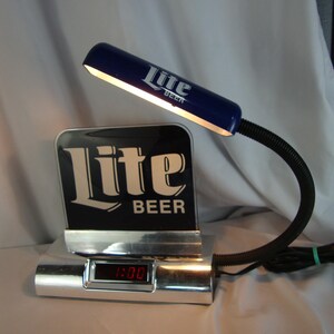 May include: A vintage desk lamp featuring the "Lite Beer" logo. The lamp has a blue shade and a flexible black neck. The base is silver with a digital clock displaying 1:00.