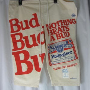 May include: Cream-colored shorts with the Budweiser logo and the phrase "NOTHING BEATS A BUD" in red. The shorts have a drawstring waist and are hanging on a black hanger.