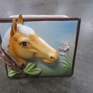 May include: A ceramic planter with a 3D image of a horse's head, a branch, and a pink flower. The planter has a brown frame and a blue background.