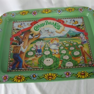 May include: A rectangular green metal tray featuring a colorful Cabbage Patch Kids design. The tray has a floral border with yellow, pink, and blue flowers. The central image depicts Cabbage Patch Kids characters in a garden setting.