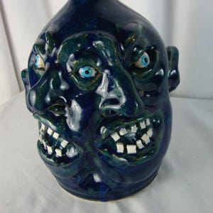 May include: A dark blue glazed ceramic jug featuring a sculpted face with three distinct eyes and two open mouths showing white square teeth. The surface has a glossy finish with deep blue and green tones, and the jug has a narrow neck opening.