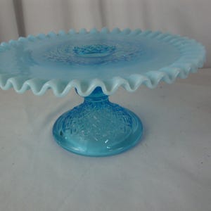 May include: A light blue glass cake stand with a ruffled edge. The round plate sits atop a pedestal base with a textured pattern. The stand is made of glass and is a vintage style.