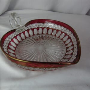 May include: Heart-shaped glass dish with a ruby red rim and gold trim. The clear glass center features a radial pattern. A small, clear handle is attached to the top. This decorative dish is suitable for serving small items.
