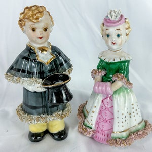May include: Two porcelain figurines of a boy and a girl. The boy wears a plaid cape and holds a black hat. The girl is in a green and pink dress with gold accents. Both have detailed facial features and are vintage collectibles.