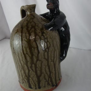 May include: A brown ceramic jug with a black bear figurine climbing on the side. The jug has a handle and a textured, dripped glaze design. The base of the jug has a red rim.