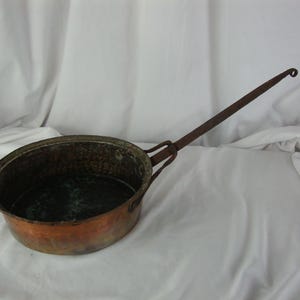 May include: A vintage copper pot with a long, curved iron handle. The pot has a hammered finish and a dark patina.