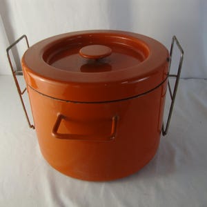 May include: A cylindrical orange metal ice bucket with a matching lid and a central knob. The container features two side handles and two additional tall, thin, rectangular metal wire frames attached to the sides that extend upwards.