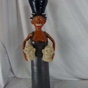 May include: A handcrafted ceramic sculpture of a person with a black hat on their head and holding two small, off-white faces. The figure has brown skin, a black dress, and a wide red smile. The sculpture is a unique piece of art.