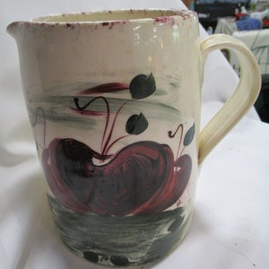 May include: A white ceramic pitcher with a handle, painted with red and green stylized fruit and leaves.