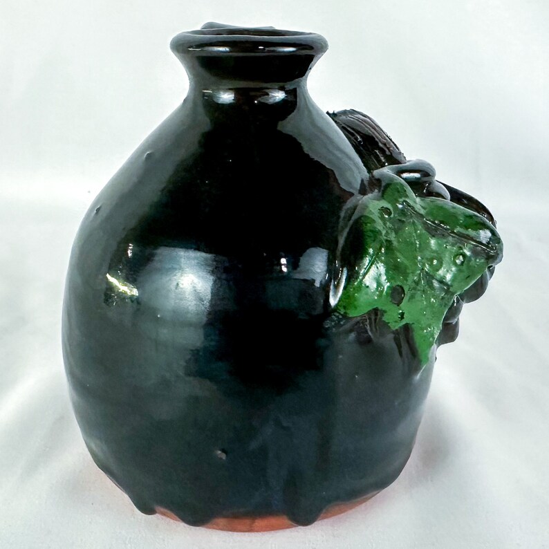 Puede incluir: Un jarr&oacute;n de cer&aacute;mica negro con un dise&ntilde;o esmaltado verde. El jarr&oacute;n tiene un cuerpo redondeado y una peque&ntilde;a abertura en la parte superior.