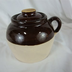 May include: A brown and white glazed ceramic crock with a lid. The crock has a handle on one side.